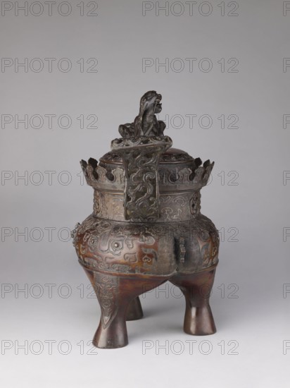 Ding-type incense burner koro with cover, Yoshida maker, Meiji period 19th/20th century, Japan, Kyoto. Incense burner with two openwork handles. Cylindrical neck with a partially openwork lattice pattern terminating in crenellation. Domed cover topped with a fo figurine holding a sphere openwork. Engraved and relief patterns over the entire surface, including karashishi/lion, taotie, komainu motifs, meander ornament and plant tendrils. Height 47.0 cm; diameter 29.0 cm., An ornate tripod vessel made of dark brown and bronze-toned metal stands on three flared legs; the rounded body and domed lid feature intricate raised patterns and decorative reliefs, with a prominent sculpted figure perched atop the lid. The upper rim has a crown-like openwork band with scalloped edges and carved motifs, and a vertical decorative panel with swirling designs rises from the rim toward the lid. The surface shows areas of patina with greenish and darker bronze hues contrasted against warmer brown highlight...