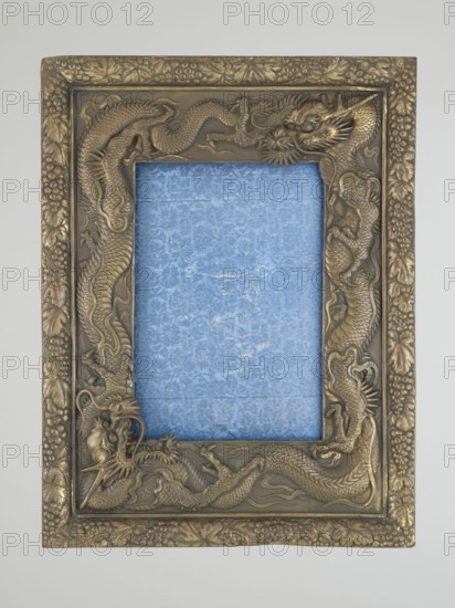 Frame with glass and raised back support covered with blue silk with floral patterns; originally for displaying. Frame decoration: relief patterns of dragons and grape clusters. 20th century, China. Materials: frame with glass and raised back support covered with silk. Height 19.5 cm; width 12.0 cm; depth 1.7 cm., A rectangular ornate picture frame with a raised dragon motif surrounding a textured inner panel, the frame showing metallic tones of gold and bronze with some darker brown shading and subtle hints of reddish highlights; the inner panel is filled with a textured blue material that has lighter and darker blue variations and a small worn area, and the outer border of the frame features carved leaf and berry decorations in matching gold and bronze hues.