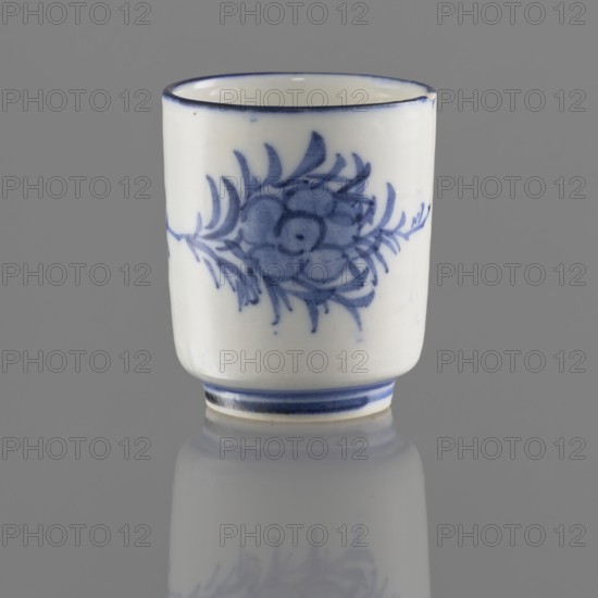 Bowl, Chinese, Qing 1644–1911, 19th/20th century. Porcelain vessel on a ring-shaped foot, height 5.5 cm, diameter 4.7 cm. Decorated with stylized floral sprigs and floral tendril ornaments. Signature in the bottom ring., A small cylindrical ceramic cup with a glossy white surface and blue painted floral decoration on its exterior, featuring a stylized flower and leafy motifs in blue, a thin blue rim at the top and blue bands near the base, sitting upright on a reflective gray surface that creates a softened mirrored reflection below; visible colors include white, several shades of blue, and gray.