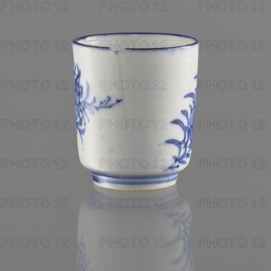 Bowl, Chinese, Qing 1644–1911, 19th/20th century. Porcelain vessel on a ring-shaped foot, height 5.5 cm, diameter 4.7 cm. Decorated with stylized floral sprigs and floral tendril ornaments. Signature in the bottom ring., A small cylindrical ceramic cup with a white base color and blue painted floral motifs on its sides, a blue rim along the top, and a blue double-line band near the foot, showing a visible crack and a small chip at the rim; the cup sits on a reflective gray surface that mirrors part of its lower section, and the background is a uniform gray.