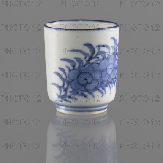 Bowl, Chinese, Qing 1644–1911, 19th/20th century. Porcelain vessel on a ring-shaped foot, height 5.5 cm, diameter 4.7 cm. Decorated with stylized floral sprigs and floral tendril ornamentation. Signature in the bottom ring., A small cylindrical ceramic cup sits upright on a reflective gray surface, showing a glossy white body decorated with a blue floral or leaf-like painted pattern wrapping around one side and a thin blue rim at the top; the cup also has a blue band near its base and a faint reflection of the painted motif on the surface below.