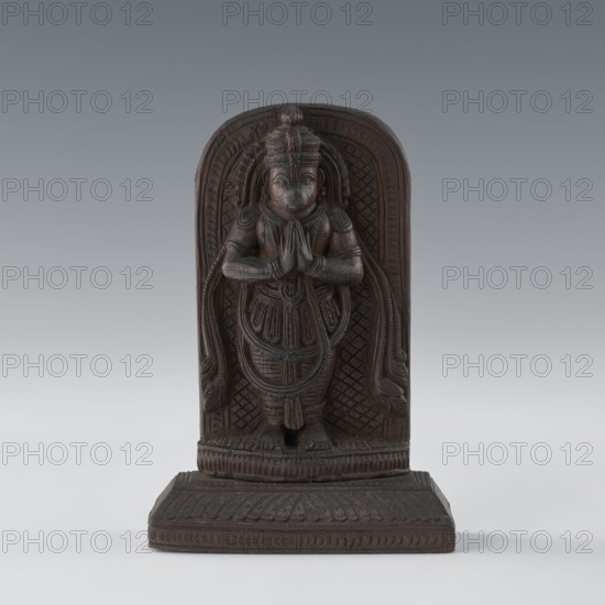 Hanuman; unknown creator; Indian, South Asian; subject: monkeys, Vaishnavism—Rama and Sita, Ramayana; Hinduism; bas-relief;, bas-relief with armlets, servants depicted; date: 1st half of the 20th century; place of origin: India; dimensions: height 9.8 cm, width 8.8 cm., A carved wooden figure in dark brown tones stands on a rectangular base with carved patterns, depicting a robed person with hands pressed together at chest level, wearing a detailed headdress and layered clothing with carved ornamental lines and textures; the figure is set against an arched carved backdrop with repeating geometric motifs and flowing carved elements at the sides, all shown on a smooth gradient background of gray and light gray.