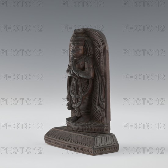 Hanuman, Indian art; South Asian/Indian culture; subject: monkeys; Vaishnavism; Rama; Sita; Ramayana; Hinduism; depiction includes armlets and servants; 1st half of the 20th century, India; bas-relief; height 9.8 cm, width 8.8 cm, A carved wooden statuette stands on a rectangular base, showing a figure in a frontal pose with hands together at chest level; the carving has detailed patterns on the figure’s clothing and headdress, layered necklaces and arm ornaments, and carved textures forming a patterned arch behind the figure. The object displays a range of brown tones in the wood, with some darker brown in recessed areas and lighter brown on raised surfaces, set against a smooth gradient background of gray and pale gray.