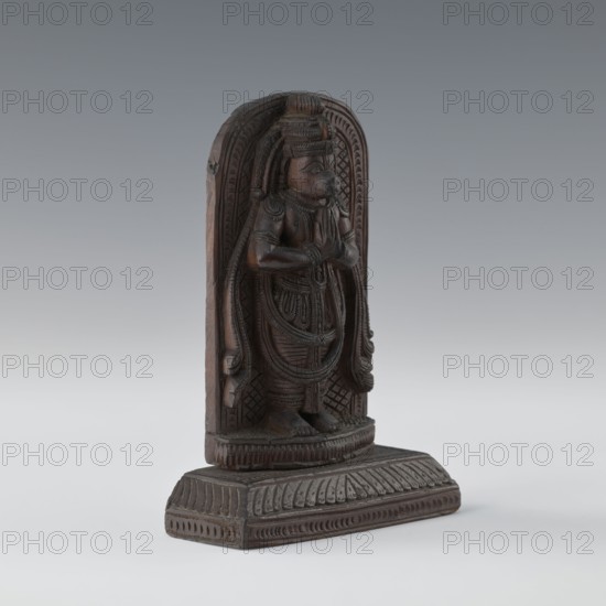Hanuman;, bas-relief; 1st half of the 20th century, India; South Asian/Indian; subjects: monkeys, Rama, Sita, Ramayana, Vaishnavism/Hinduism; depiction includes armlets and servants; height 9.8 cm, width 8.8 cm., A carved wooden standing figure mounted on a detailed rectangular base, carved in dark brown wood with visible grain and polished areas; the figure is framed by an arched backing with carved geometric and linear patterns, and the figure itself wears layered draped garments and ornate jewelry indicated by carved lines and raised bands, with hands held together at chest level, a headdress with vertical ridges, and stylized hair or adornments falling at the sides; the base and arch feature repeating carved motifs and borders, and the overall colors visible are dark brown wood tones with lighter brown highlights and subtle blackened shadows against a neutral gray background.