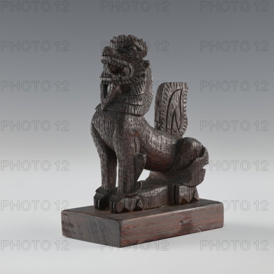 Lion, Indian/South Asian, 1st half of the 20th century, figurine, animals, claws, height 13.2 cm, width 9.7 cm, possible origin India or Sri Lanka., A carved wooden figure of an animal-like creature with a textured surface sits on a rectangular wooden base; the creature has a raised head with an open mouth and visible carved teeth, a mane or layered neck, a curved tail that rises behind its body, and four legs with detailed paws. The carving shows a rough, chiseled finish with visible tool marks. The colors visible are dark brown and reddish-brown on the figure and base, with lighter gray and pale gray in the neutral background.
