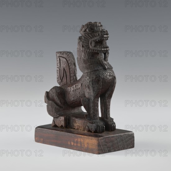Lion;, figurine; Indian/South Asian culture; animals; claws; 1st half of the 20th century; India, / Sri Lanka ; height 13.2 cm; width 9.7 cm, A carved wooden sculpture of a stylized animal with a raised head and open mouth sits on a rectangular wooden base; the figure has a textured surface with visible carving marks, a mane-like neck, pronounced front legs with carved paws, a curled tail, and a vertical back panel rising behind the body. The visible colors are dark brown and lighter brown variations on the wood, with grey background tones.
