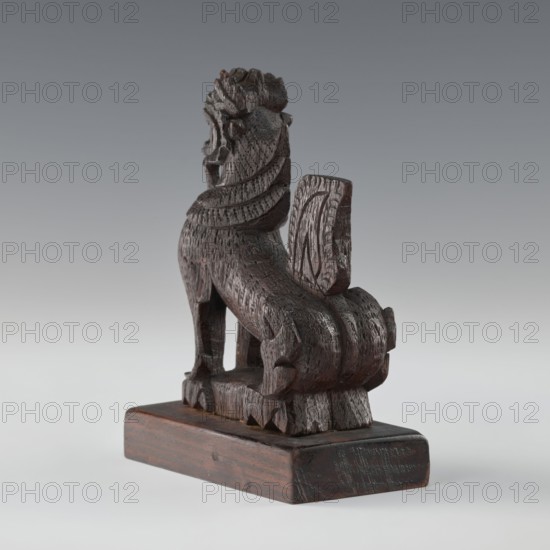 Lion;, ; Indian/South Asian; animals; claws; 1st half of the 20th century; India, Sri Lanka ; figurine; height 13.2 cm; width 9.7 cm., A carved dark brown wooden figure of an animal-like creature sits on a simple rectangular wooden base, showing textured carved details across its body and mane, with a raised tail element behind it; the object has visible wood grain and tool marks and presents a glossy patina in places, while the background is a smooth gradient of gray transitioning from darker at the top to lighter toward the bottom.