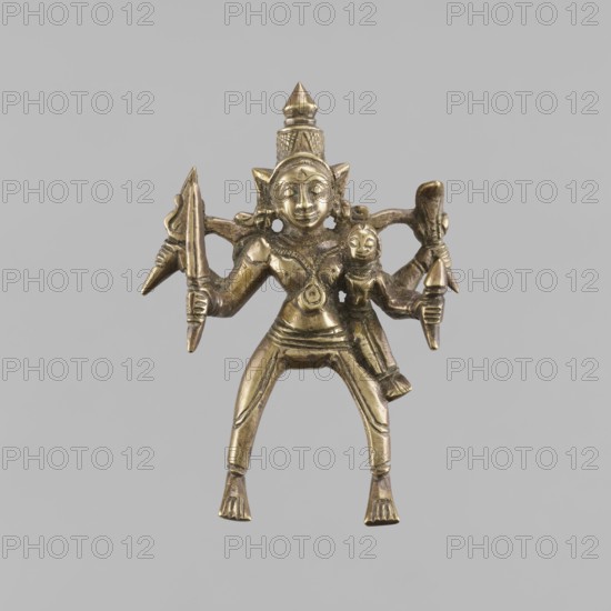 Four-Armed Divine Warrior Shiva as Khandoba?, Indian, Shaivism, Hinduism, 1st half of the 20th century. Bronze figurine, height 11.5 cm, width 8.0 cm. Depicts a four-armed divine warrior with swords, trident, crown/tiara, snakes; themes of violence, warriors, gods, couples and marriages., A small metallic figurine in a brass or gold tone depicting a standing humanoid figure with a conical headdress and pointed ears, shown with a broad torso, bent legs, and flat feet; the figure has multiple arms holding various object-like shapes and a smaller human-like figure attached at one side, with carved facial features, decorative chest and waist details, and textured elements on the headdress and limbs, all set against a plain light gray background.