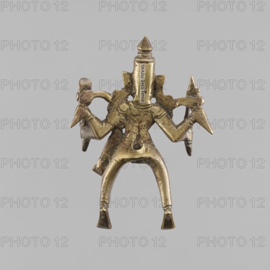 Four-Armed Divine Warrior Shiva as Khandoba?, Indian, Shaivism/Hinduism, 1st half of the 20th century. Figurine, height 11.5 cm, width 8.0 cm. Subjects/features: four-armed divine warrior, gods, warriors, swords, crowns/tiaras, tridents, snakes; themes of violence, couples, marriages., Indian art., A small metallic figurine with a worn golden-brass color is shown against a neutral gray background, depicting a stylized humanoid shape with a tall pointed headpiece, broad shoulders, and a torso with textured surface marks. The figure has a pair of bent legs forming an arch and flat, trapezoidal feet, and each arm extends outward holding angular, pointed objects with darker metallic tips. There are decorative protrusions on the shoulders and a few small perforations through the body, with subtle surface scratches and patina variations in gold, brass, and darker brown tones.
