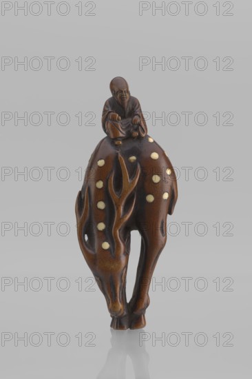 Netsuke in the shape of Fukurokuju riding a deer. The deer has all its legs put together so that their hooves touch each other; the lowered head touches the front hooves, causing the animal's body to be curved into the shape of an irregular oval, on top of which sits a small figurine of Fukurokuju, the deity of longevity. Unsigned. Mid 19th century Edo period, Japan. Sculpture, netsuke katabori. Height 9.8 cm; width 4.2 cm., A small carved figurine in brown tones with a glossy finish depicts a seated human figure atop an animal-like form; the human figure is darker brown and shows facial features and folded clothing with hands resting forward. The animal-like form is a lighter warm brown with carved flowing shapes resembling antlers or branches along its sides and several pale circular inlays scattered across its back and sides. The piece stands on narrow legs that taper to rounded feet and casts a soft reflection on a smooth light gray surface. The background is a uniform pale gray.