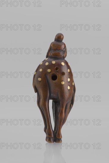 Netsuke in the shape of Fukurokuju riding a deer. The deer has all its legs put together so that their hooves touch each other; the lowered head touches the front hooves, curving the body into an irregular oval, on top of which sits a small figurine of Fukurokuju, the deity of longevity. Unsigned. Mid 19th century Edo period, Japan. Sculpture, netsuke katabori. Height 9.8 cm, width 4.2 cm., A small carved wooden figure of a person seated on the back of an animal, shown from the rear; the figure and animal are a polished brown wood with a warm, reddish tone, and the animal's rounded back is decorated with a group of light cream circular inlays; there are a few circular holes visible on the animal's back, and the legs of the animal are elongated and slender, ending in rounded hooves; the surface shows smooth carved contours and subtle darker grain variations, and the whole object sits against a plain light gray background with a faint reflection beneath it.