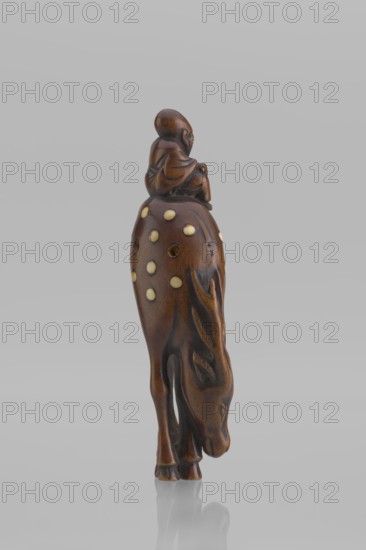 Netsuke in the shape of Fukurokuju riding a deer. The deer has all its legs put together so that their hooves touch each other; the lowered head touches the front hooves, causing the animal's body to be curved into the shape of an irregular oval, on top of which sits a small figurine of Fukurokuju, the deity of longevity. Unsigned. Mid 19th century Edo period, Japan. Sculpture, netsuke katabori. Height 9.8 cm; width 4.2 cm., A carved brown wooden figurine depicting a person seated on the back of an animal with its head lowered; the wood shows darker and lighter brown tones with smooth polished surfaces, and the animal’s back is decorated with a group of pale circular inlays.