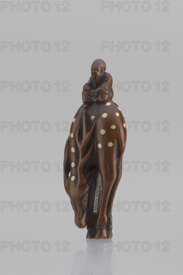 Netsuke in the shape of Fukurokuju riding a deer. The deer has all its legs put together so that their hooves touch each other; the lowered head touches the front hooves, causing the animal's body to be curved into the shape of an irregular oval, on top of which sits a small figurine of Fukurokuju, the deity of longevity. Unsigned. Mid-19th century Edo period, Japan. Sculpture, netsuke katabori. Height 9.8 cm; width 4.2 cm., A small carved brown wooden figure shows a person seated atop a deer-like animal. The person appears to wear flowing robes and has a rounded head and facial features carved in a darker brown shade. The animal has a lowered head and slender legs, with carved antler-like ridges and a pattern of light-colored circular inlays on its back and sides. The overall colors visible are brown, darker brown accents, and pale cream inlays, set against a neutral light gray background.