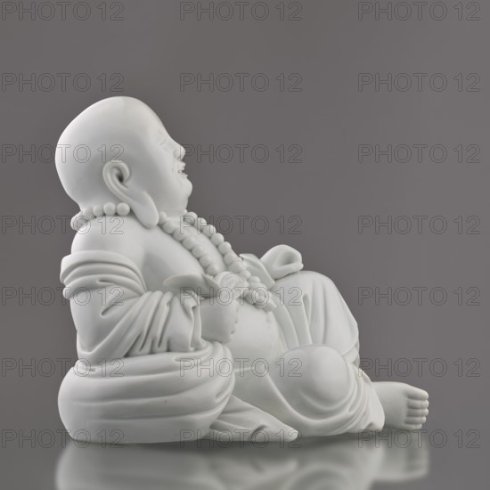 Budai Laughing Buddha, Chinese, Qing dynasty 1644–1911, 19th century, China, Fujian; subject: Budai Buddha of the Future, Maitreya, Milefo; material: figurine; dimensions: height 30.4 cm, width 21.6 cm, weight 3 kg., A smooth white ceramic statue of a seated, smiling figure wearing beaded necklaces and flowing robes, shown in profile against a solid gray background; the statue rests on a rounded base and casts a subtle reflection on the surface beneath it, with visible details in the folds of the fabric and the rounded features of the face, hands, and feet.
