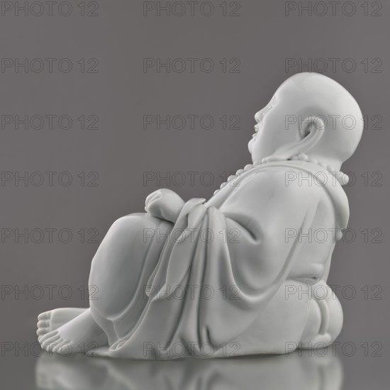 Budai Laughing Buddha, from Ignacy Jan Paderewski, Chinese, Qing dynasty 1644–1911, Budai iconography representing the Buddha of the Future / Maitreya Milefo, 19th century, Fujian, China; figurine; height 30.4 cm, width 21.6 cm, weight 3 kg., A smooth white sculpted figure seated in a relaxed pose, shown in profile against a neutral gray background, with rounded facial features and a serene expression, a rounded bald head, a beaded necklace around the neck, draped flowing robes with soft folds and layered fabric, hands resting near the torso, bare feet visible with detailed toes, a rounded belly, and a subtle reflective surface beneath that mirrors parts of the base; the visible colors are white and shades of gray.