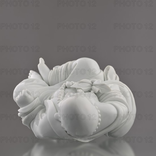 Budai Laughing Buddha, Chinese, Qing dynasty 1644–1911, 19th century, Fujian, figurine representing Budai / Maitreya Buddha of the Future, height 30.4 cm, width 21.6 cm, weight 3 kg., A glossy white ceramic figurine of a reclining, chubby childlike figure wearing draped clothing and a beaded necklace, shown against a smooth gray background; the figure has rounded limbs, a bald head, and detailed folds in the fabric, with highlights and reflections on the shiny surface and a soft reflection of the piece visible beneath it.