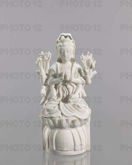 Guanyin on a lotus throne, Chinese, Blanc de Chine porcelain, 18th century, Fujian, China; sculpture of Guanyin goddess in Chinese Buddhism, height 27.0 cm, width 14.3 cm., A white glazed porcelain figure of a seated multi-armed deity on a rounded pedestal, set against a smooth light gray background; the figure shows a calm facial expression, a detailed headdress, draped robes with flowing folds, several arms arranged around the body holding stylized lotus-shaped elements, and a decorative base with petal-like and beaded motifs, all rendered in glossy white with subtle shadows and highlights.
