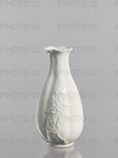 Vase, gift; Chinese; Qing dynasty 1644–1911; peaches iconography; decorative vessel; 1690–1710, China; height 17.5 cm, diameter 8.4 cm., A white ceramic vase with a glossy finish stands against a plain light gray background; the vase has a flared, ruffled rim and a gently tapered, rounded body with raised floral decorations on one side, and the surface shows subtle crackle texture and soft reflections.