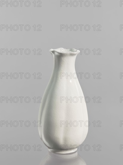 Vase, Chinese, Qing dynasty 1644–1911, decorated with peaches, decorative vessel, 1690–1710, China; vessel; height 17.5 cm, diameter 8.4 cm., A single white ceramic vase with a glossy finish and a gently flared, wavy rim is centered against a smooth light gray background, showing subtle crackle texture and soft reflections on its curved surface, with a faint reflection of the vase visible on the reflective surface below; visible colors include white and shades of gray.