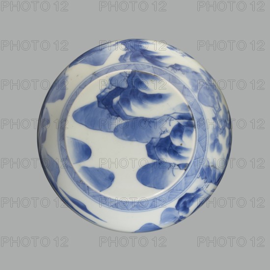 Storage Vessel with Lid, Zôshuntei Sanpo 1841–1878. Storage pot, blue-and-white porcelain, Japanese; pear-shaped vessel with flat cylindrical lid. Decorated in a Chinese-style landscape: an old man resting alone under a pine on a rocky cliff overlooking a vast body of water, with a picturesque mountainous landscape, waterfall, lofty peaks, and distant boat sails. Lower base: wide ornamental band of three vertical and two crossed lines. Neck and lower edge of lid: ornamental bands of diagonal lines forming triangular forms. Lid decorated with a rock motif. Decoration modeled on Chinese Southern School painting, related to the Japanese bunjinga nanga movement; free-form painting with precise, masterful lines. Cobalt in a bright, pure shade in several tones. Underglaze cobalt signature on base: Zoshuntei Sampo zo made by Zoshuntei Sampo. c. 1842–1878. Arita, Japan. Height 21.8 cm; diameter 15.3 cm., A round ceramic plate shown from above with a white background and blue painted floral-lik...