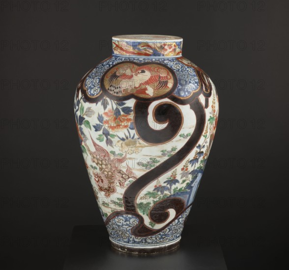 Vase tsubo, Japanese, decorated with butterflies, birds, phoenixes hoo, karashishi, paulownia, chrysanthemums, peonies, falcons, and plant tendrils in kachôga style; colored porcelain Imari, Arita, ca. 1720–1740 Edo Period, 1603–1868; vessel, height 63.0 cm., A large ceramic vase with a rounded body and narrow neck sits against a dark background, decorated with a bold dark brown curving scroll motif and ornate panels containing stylized birds and floral designs; visible colors include white, dark brown, blue, red, green, orange, and gold, with patterned blue borders near the base and neck, painted leafy branches and blossoms in green, blue, and orange, and areas of intricate red and gold detailing on the upper panels, all resting on a dark plinth.