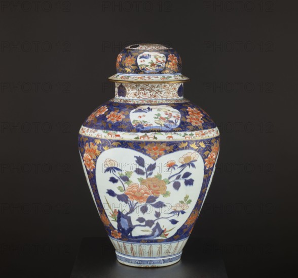 Vase tsubo with cover, Japanese, Imari ware, Edo period 1603–1868, decorated with plum trees, meander ornament, plant tendrils, bamboos, peonies, pine trees, and genre scenes; Arita, Japan; height 68.5 cm, diameter 45.0 cm; dated 1690–1730; vessel., A lidded ceramic vase with a rounded body and narrow base sitting against a dark background; the vase shows a decorative palette of deep blue, white, orange, green, and gold, with several floral motifs and leafy branches painted on its main panels, a few heart-shaped white cartouches containing flower scenes, and bands of intricate floral and foliate patterns around the shoulder and neck.