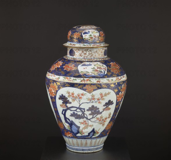 Vase tsubo with cover, Japanese, Imari style, Edo period 1603–1868, decorated with plum trees, bamboos, peonies, pine trees, plant tendrils, meander ornament, and genre scenes; Arita, Japan; 1690–1730; vessel; height 68.5 cm, diameter 45.0 cm., A lidded ceramic vase sits against a dark background, showing a bulbous body narrowing to a short foot and topped with a rounded lid; the vase displays a decorative palette of deep blue, white, orange, gold, green, and touches of red, with large white cartouche-shaped panels featuring a floral branch motif in dark blue and orange, surrounded by deep blue fields filled with stylized orange flowers and gold foliate patterns, a band with small painted scenes around the shoulder, a flared rim with intricate gold and blue ornament, and a foot rim decorated with blue geometric stripes.