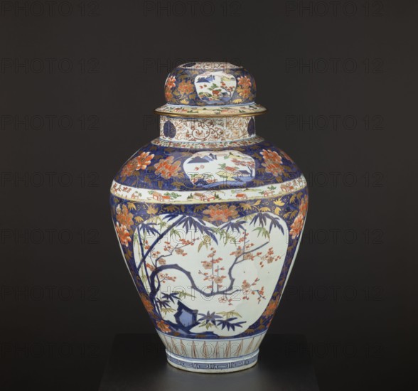Vase tsubo with cover, Japanese, Imari style, Edo period 1603–1868, circa 1690–1730, Arita, Japan. Porcelain vessel, height 68.5 cm, diameter 45.0 cm. Decorated with floral motifs including plum trees, bamboos, peonies, pine trees, plant tendrils, meander ornament, and genre scenes., A lidded ceramic vase with a rounded body and a narrower neck displayed against a dark background; the vase shows a palette of deep blue, white, orange, green, gold, and touches of brown. The lid and upper shoulder feature deep blue areas decorated with orange floral motifs and gold scrolling, and there are panels of white with painted landscape motifs. The main body has a large white panel bordered by deep blue containing a painted scene of dark blue branches with orange blossoms and green leaves, along with additional floral and bamboo motifs around it. A band of white with small painted house-like shapes encircles the vase near the top, and gold decorative elements appear in the blue areas. The vase sit...