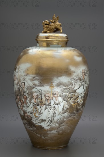 Vase with cover, Kanzan, Denshichi 1821–1890 maker, Japanese, Meiji period 1868–1912, late 19th century; ceramics; ovoid with a gently upward-flaring belly terminating in a narrow short collar; decorated with rakan, lions/karashishi iconography; vessel, height 61 cm, diameter 34.5 cm., A large lidded ceramic vase with a rounded body and a domed lid topped by a sculpted animal figure in metallic gold sits against a neutral gray background; the vase surface shows broad areas of metallic gold and ivory white with painted scenes of robed figures rendered in muted tones of white, beige, brown, and gray, accented with gold detailing; the lid and neck feature patterned gold decoration, and the sculpted finial is gold; the painted figures wear flowing white robes and appear among stylized cloud-like forms in shades of gray and beige, with additional small areas of gilded ornamentation and subtle darker line work defining faces and garments.