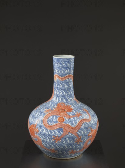 Vase-bottle, Chinese, Qing dynasty Qianlong, 1736–1795, 18th century. Blue-and-white porcelain vessel with bulbous belly, slightly flattened at the top; decoration of dragons with a pearl amid waves/sea waves and pearls. Signed. Height 54 cm; diameter 46 cm., A bulbous ceramic vase with a long narrow neck sits against a dark gray background, decorated with a repeating wave pattern in blue and white and featuring a few large stylized dragons in orange wrapping around the body and neck; the vase rests on a dark surface and shows glossy ceramic highlights and subtle shadowing.