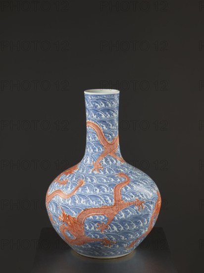 Vase-bottle, Chinese, Qing dynasty Qianlong, 1736–1795, 18th century. Blue-and-white porcelain vessel, bulbous belly slightly flattened at the top; decorated with dragons, dragon with a pearl, waves/sea waves, and pearls. Signed. Height 54 cm, diameter 46 cm., A rounded ceramic vase with a long narrow neck sits against a dark background, decorated overall with a repeating wave pattern in blue and white and overlaid with sinuous dragon forms in orange-red that wrap around the body and extend onto the neck, the vase showing glossy surfaces and smooth contours with the blue, white, and orange-red colors prominent.