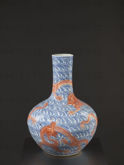 Vase-bottle, 18th century China, Qing dynasty, Qianlong period 1736–1795. Blue-and-white porcelain with painted dragons, dragon with a pearl, waves and sea waves, pearls; bulbous belly slightly flattened at the top. Signed. Height 54 cm, diameter 46 cm., A rounded ceramic vase with a long narrow neck sits against a dark background, decorated with a repeating wave pattern in blue and white across its surface and overlaid with several sinuous dragon figures in orange and reddish tones; the dragons have detailed scales and claws, and the base of the vase shows a slightly warmer, beige rim.