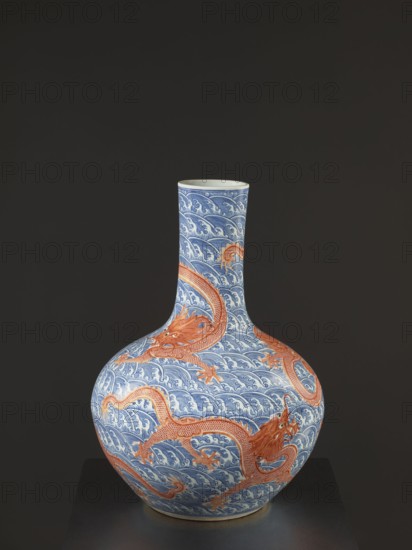 Vase-bottle, Chinese, Qing dynasty, Qianlong period 1736–1795, 18th century. Blue-and-white porcelain; bulbous belly slightly flattened at top; decorated with dragons, dragon with a pearl, waves and sea waves, pearls. Signed. Height 54 cm, diameter 46 cm., A rounded ceramic vase with a long narrow neck sits against a dark background, decorated in blue and white wave patterns covering its surface and accented by a few large orange-red dragon motifs that wrap around the body and neck; the top rim appears pale, and the base shows a touch of warm beige.