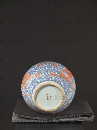 Vase-bottle, Qing dynasty, Qianlong period 1736–1795, 18th century China. Blue-and-white porcelain vessel with bulbous belly slightly flattened at the top; decoration of dragons including dragon with a pearl, waves/sea waves, and pearls. Signed. Height 54 cm, diameter 46 cm., A rounded ceramic vase with decorative patterns rests on a folded fabric cushion on a dark surface against a dark background; the vase shows a design of flowing wave-like motifs in blue with dragon figures in orange wrapping around its curved body, and the base rim shows a natural beige ceramic edge with an inner pale area; the cushion beneath appears gray with stitched seams and a slightly textured surface, and the overall scene includes colors such as blue, orange, beige, gray, pale off-white, and dark brown or black in the background.