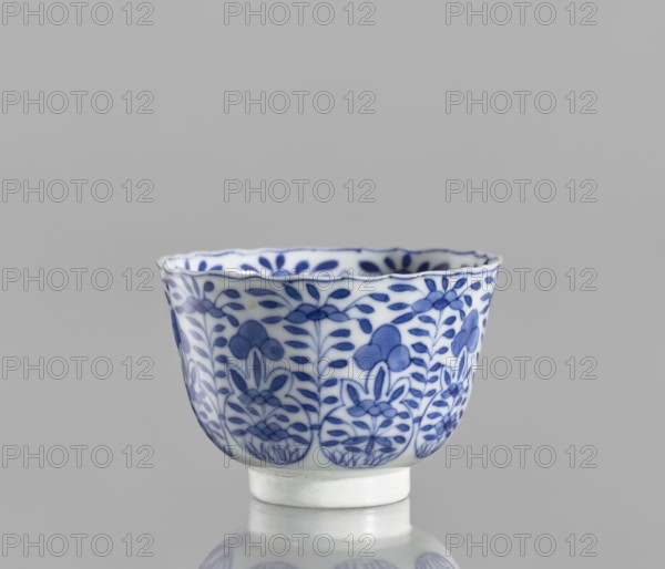 Bowl, Chinese blue-and-white porcelain, ceramics; floral and plant ornament decoration; conical with serrated rim; Kangxi period 1662–1722, Qing dynasty 1644–1911; China; vessel; height 5.6 cm, diameter 7.9 cm., A small porcelain cup with a scalloped rim sits against a plain gray background, decorated with a floral pattern in blue on a white ground; the design shows a repeating motif of stylized flowers and leaves wrapping around the exterior, and the cup rests on a short white foot that is reflected faintly on the glossy surface below.