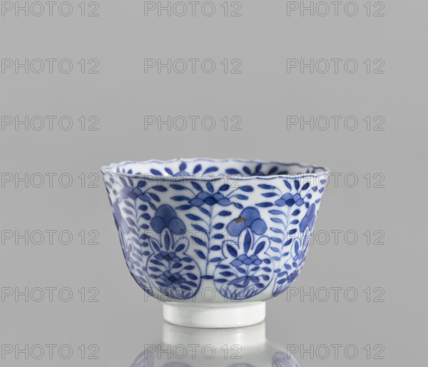 Bowl, Chinese blue-and-white porcelain, Chinese culture, ceramics; floral and plant ornament decoration; Kangxi period 1662–1722, Qing dynasty 1644–1911; conical with serrated rim; 1662–1722; China; vessel; height 5.6 cm, diameter 7.9 cm, A small porcelain cup with a scalloped rim sits against a plain gray background, decorated all around with a repeating blue floral and leaf pattern on a white ground; the blue decoration includes darker and lighter blue shades forming stylized blossoms and leafy vines, and the cup rests on a short white foot with a slight reflection visible beneath it.