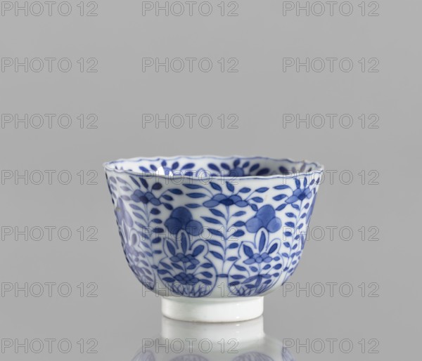 Bowl, Chinese, blue-and-white porcelain, ceramics; floral and plant ornament decoration; conical with serrated rim; Kangxi period 1662–1722, Qing dynasty 1644–1911; China; vessel; height 5.6 cm, diameter 7.9 cm, A small white ceramic cup with blue floral patterns around its exterior sits against a neutral gray background, showing a scalloped rim and a short cylindrical foot; the blue decoration features stylized leaves and flower motifs arranged in repeating vertical bands, and the cup casts a faint reflection on the surface below, with visible colors including white, blue, gray, and a slight off-white on the foot.