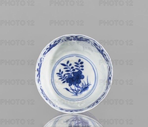 Bowl, Chinese blue-and-white porcelain, floral and plant ornament decorations, conical with serrated rim, Kangxi period 1662–1722, Qing dynasty 1644–1911, China; vessel, height 5.6 cm, diameter 7.9 cm., A round ceramic bowl with a white base and blue painted decoration, shown from above against a light gray background; the interior features a blue floral motif surrounded by concentric circular bands and a decorative blue border along the rim; the rim has a scalloped edge and the glaze shows subtle reflections and highlights; the bowl and its painted design are reflected faintly on the surface below.