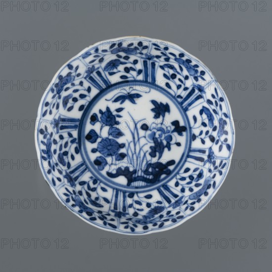 Bowl with a serrated edge, Chinese, Qing dynasty 1644–1911, Xuande style 1426–1435, blue-and-white porcelain; decoration: inside mirror with flowers, side walls with segments of plant decoration—peonies, lotuses, rocks; backdated signatures; ceramics vessel, height 3.3 cm, diameter 9 cm; dated 1662–1722., A round ceramic plate with a scalloped edge sits against a gray background; the plate is white with intricate blue painted designs, featuring a central circular medallion filled with floral motifs and slender leaves, surrounded by a patterned band of alternating vegetal elements and fan-like segments, and an outer rim decorated with repeating leafy sprigs and small blossoms in shades of deep blue and navy on the white surface.