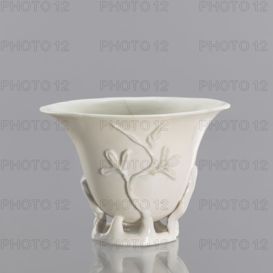 A bowl shaped like rhinoceros-horn carving, Chinese, Qing dynasty 1644–1911, blanc de Chine porcelain with plant ornament and plastic decoration imitating rhinoceros horn, 19th century, Fujian, vessel, height 19 cm, diameter 9.2 cm, A white ceramic cup with a flared rim sits against a pale gray background, showing a smooth glossy surface and a raised branch-and-leaf relief on its side; the cup narrows toward a decorative openwork foot formed by a group of curved supports, and subtle shadows and reflections appear on the surface beneath it.