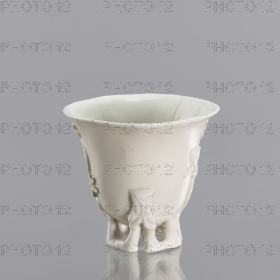 Bowl shaped to resemble one carved in rhinoceros horn; maker, Qing dynasty 1644–1911, 19th century; blanc de Chine porcelain with plant ornament and modeled plastic decoration imitating rhinoceros horn carving; Chinese, Fujian; vessel, height 19 cm, diameter 9.2 cm., A white ceramic cup with a flared rim sits against a gray background, showing a smooth glossy surface with subtle decorative relief on its sides and a few fine hairline markings, supported by a group of small sculpted feet that create a reflected shape on the shiny surface beneath; visible colors include white and gray.
