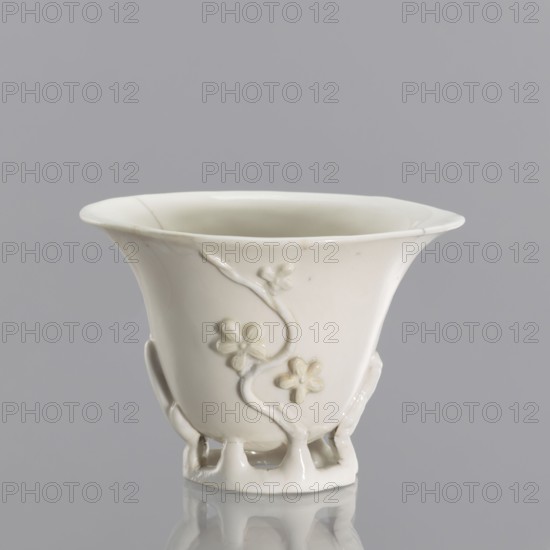 A bowl in a shape reminiscent of a bowl carved in rhinoceros horn; creator, Chinese; Qing dynasty 1644–1911; 19th century; blanc de Chine porcelain with plant ornament and plastic decoration imitating rhinoceros horn carving; from Fujian, China; vessel; height 19 cm, diameter 9.2 cm., A white ceramic cup with a flared rim and a glossy finish sits against a gray background, showing a decorative raised floral vine motif on its exterior and an open lattice-like foot, with subtle shadows and reflections visible on the surface.