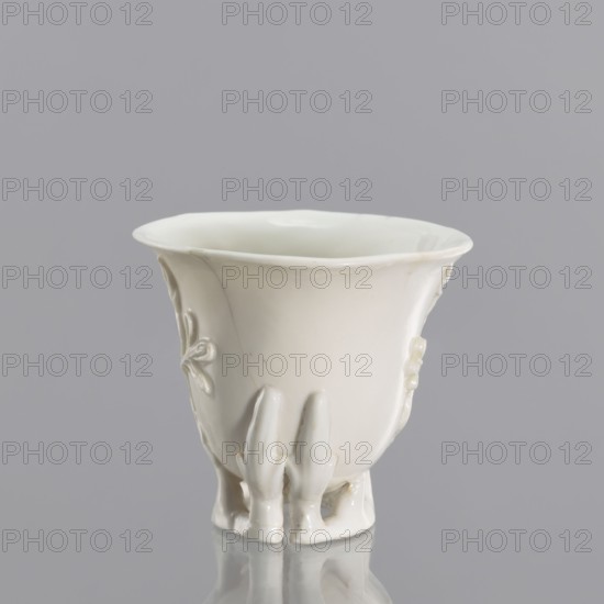 A bowl shaped to resemble rhinoceros horn, Chinese, Qing dynasty 1644–1911, 19th century; blanc de Chine porcelain with plant ornament and plastic appliqué decoration; from Fujian, China; vessel, height 19 cm, diameter 9.2 cm., A glossy white ceramic cup with a flared rim sits upright on a reflective surface against a smooth gray background; the cup has a rounded body with subtle raised decorative motifs on its sides and three short sculpted feet at the base, and visible highlights and soft shadows emphasize its curved form and glossy texture.
