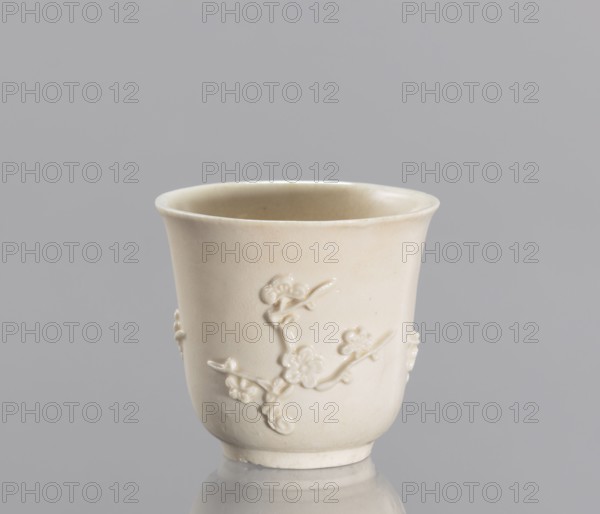 Bowl, plum trees, porcelain blanc de Chine, Chinese art, Chinese culture, decorative vessel with applied/plastic decoration, 19th century China, vessel, height 7 cm, diameter 7.2 cm, A single pale cream-colored ceramic cup sits against a gray background, showing a glossy finish and a slightly flared rim; the cup's surface features a raised floral branch decoration wrapping around its exterior, with the relief petals and stems rendered in the same cream tone, and the cup casts a subtle reflection on the smooth surface beneath it.
