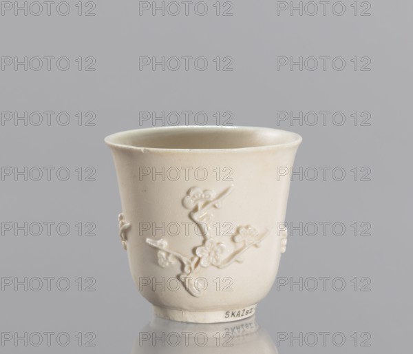 Bowl, decorated with plum trees, porcelain blanc de Chine, 19th century China; ceramic decorative vessel with plastic relief decoration; height 7 cm, diameter 7.2 cm., A small off-white ceramic cup with a smooth rounded rim and a slightly flared top, showing a raised floral branch motif wrapping around its exterior; the cup has a matte, creamy beige surface with subtle variations in tone and a few small imperfections in the glaze, and it sits on a reflective surface against a light gray background.