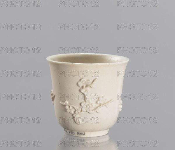 Bowl, decorated with plum trees, porcelain blanc de Chine, Chinese, plastic decoration, 19th century China, vessel, height 7 cm, diameter 7.2 cm, An off-white ceramic cup with a slightly flared rim sits against a gray background; the cup has a smooth glazed surface with a raised floral branch motif wrapping around its body, showing sculpted flowers and stems in the same off-white color, and a subtle reflective sheen at the base where it rests on a reflective surface.