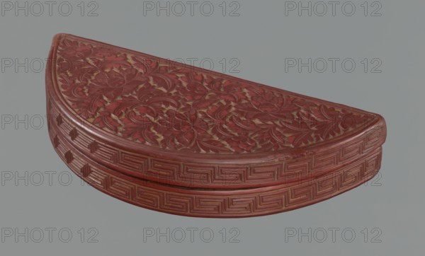 Box with a lid, semi-circular container decorated on the lid with a dense pattern of peony flowers and leaves; rim decorated with a Chinese meander motif. Materials/ornament: leiwen meander, peonies, lemons. Chinese art, Ming dynasty 1368–1644, 17th century, China. Height 4.4 cm, width 24.0 cm, depth 10.7 cm., A carved semicircular object with an ornate floral surface and geometric patterned sides, predominantly red with visible gold or yellow accents in recessed areas, set against a plain gray background.