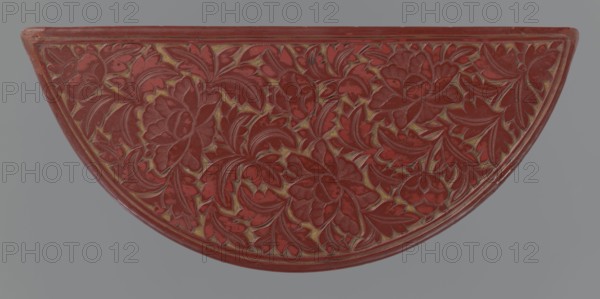Box with a lid, Chinese, Ming 1368–1644, 17th century. Semi-circular box decorated on the lid with a dense pattern of peony flowers and leaves; rim decorated with a Chinese meander motif. Materials/technique suggested by form: container. Dimensions: height 4.4 cm, width 24.0 cm, depth 10.7 cm. Subjects/ornament: peonies, leiwen meander, lemons., A semicircular object with an ornate carved surface covered in a floral relief pattern, showing a group of overlapping stylized flowers and leaves with curved petals and lobed foliage; the carved areas are predominantly red with worn highlights showing a goldish tone in the recesses, and the background around the object is a muted gray.