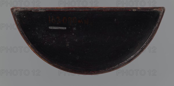Box with a lid;, gift; Chinese art; Chinese; Ming 1368–1644; boxes; peonies; meander ornament; leiwen ornament; lemons. Semi-circular box decorated on the lid with a dense pattern composed of peony flowers and leaves. The rim is decorated with a Chinese meander motif. 17th century; China; container. Height 4.4 cm, width 24.0 cm, depth 10.7 cm., A semicircular object with a dark, glossy surface and a worn reddish-brown rim sits against a neutral gray background; the dark surface shows small scattered lighter marks and scuffs, and the rim has areas of chipped paint revealing lighter wood tones, with overall colors including black, dark brown, reddish-brown, gray, and a few small pale flecks.