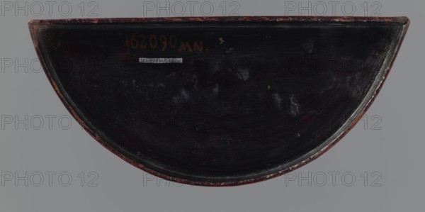 Box with a lid, semi-circular container decorated on the lid with a dense pattern of peony flowers and leaves; rim decorated with a Chinese meander motif. Materials/technique and, Chinese, Ming dynasty 1368–1644, 17th century. Motifs: peonies, leiwen meander ornament, lemons. Dimensions: height 4.4 cm, width 24.0 cm, depth 10.7 cm., A semicircular object with a smooth, dark surface that appears black with subtle reddish undertones and faint glossy highlights, bordered by a worn rim showing a mix of dark red and brown tones; the object rests against a uniform gray background and displays a few scuff marks and smudges on its face.